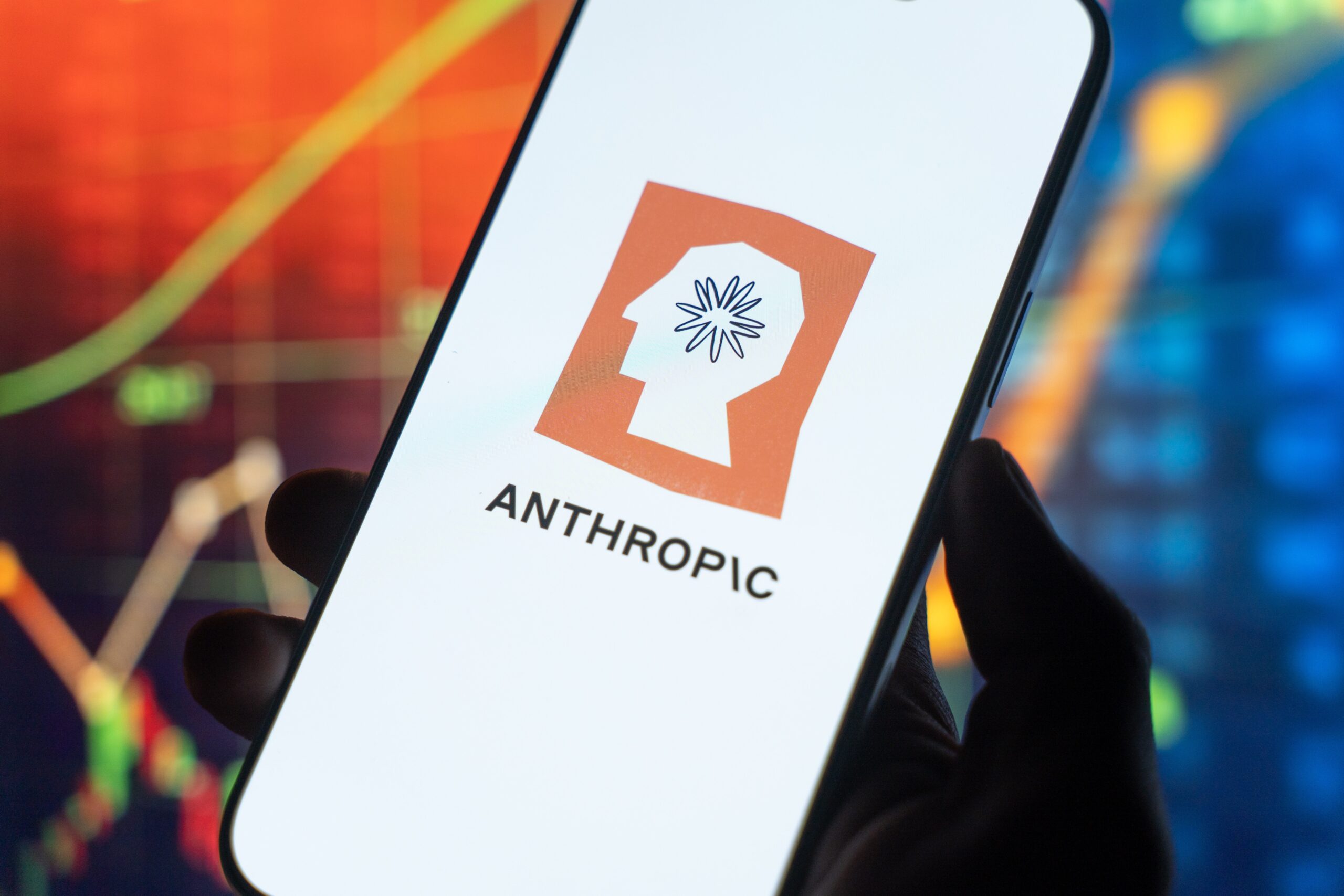 โลโก้ Anthropic บนหน้าจอสมาร์ทโฟน วางอยู่หน้าฉากหลังกราฟราคาหุ้นที่สื่อถึงความกังวลของนักลงทุน